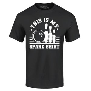 Può includere: Maglietta nera con una grafica bianca di una palla da bowling e birilli. Il testo "This is my spare shirt" è stampato sulla maglietta.