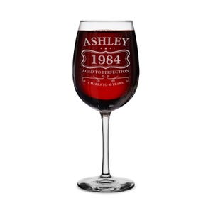 May include: A clear glass wine glass with a white inscription that reads "Ashley 1984 Aged to Perfection Cheers to 40 Years". The glass is filled with red wine.