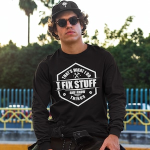 May include: Black long-sleeve shirt with a white graphic that says "That's What I Do I Fix Stuff And I Know Things". The graphic is in a hexagonal shape with a wrench and screwdriver design.