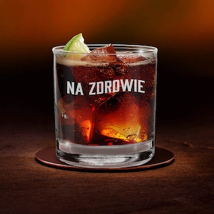 Puede incluir: Un vaso de vidrio transparente con un líquido marrón oscuro y cubitos de hielo. Una rodaja de lima se asienta en el borde del vaso. El vaso tiene el texto "NA ZDROWIE" impreso en él.