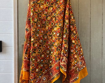 May include: A yellow, red and green embroidered shawl with mirror embellishments. The shawl is draped over a hanger and has a geometric pattern.