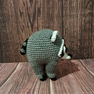 May include: A round, crocheted raccoon plush toy featuring a gray body, a black and white striped tail, and a black and white face mask. The small stuffed animal is crafted with visible yarn stitches and has a compact, spherical shape with four short legs.