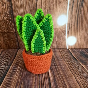 May include: A crocheted succulent plant in a brown pot. The plant has green leaves and is sitting on a wooden surface.