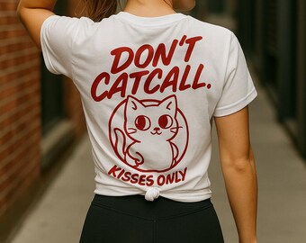 May include: White t-shirt with the text "DON'T CATCALL" in red, along with a cartoon cat illustration and the words "KISSES ONLY." The shirt is knotted at the waist. The person is wearing black shorts.