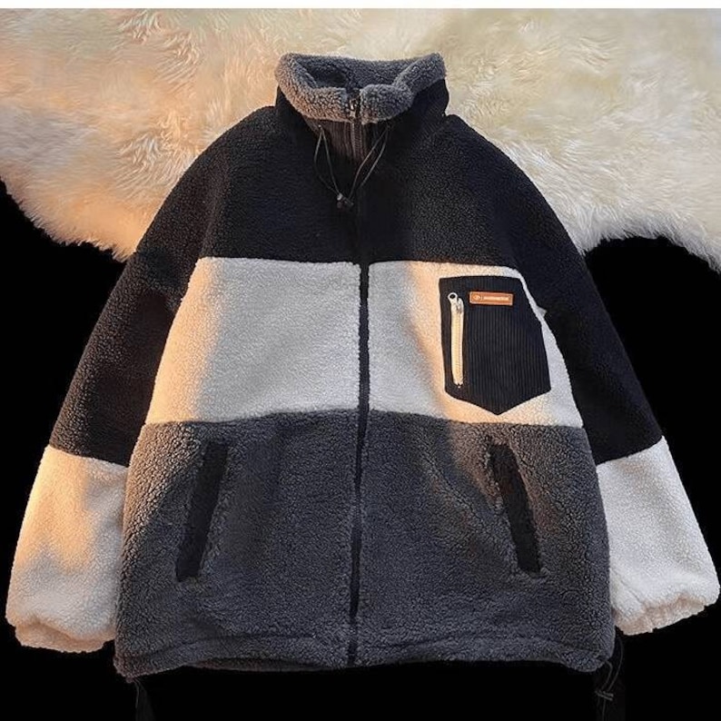 May include: A black, white, and gray fleece jacket with a zipper closure and a small pocket on the right side. The pocket has a zipper and a brown label with text.