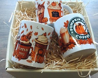 Puede incluir: Conjunto de cuatro tazas de cerámica blanca con diseños de otoño. Las tazas presentan imágenes de cafés con especias de calabaza, café y hojas de otoño. Las tazas están dispuestas en una cesta llena de papel triturado.