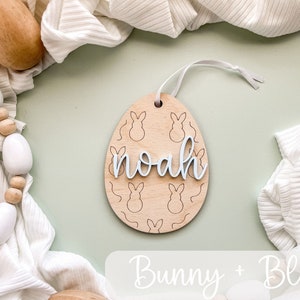 May include: A wooden Easter egg ornament with a bunny pattern and the name "noah" written in white. The ornament is hanging from a white ribbon and is surrounded by white fabric and wooden beads.