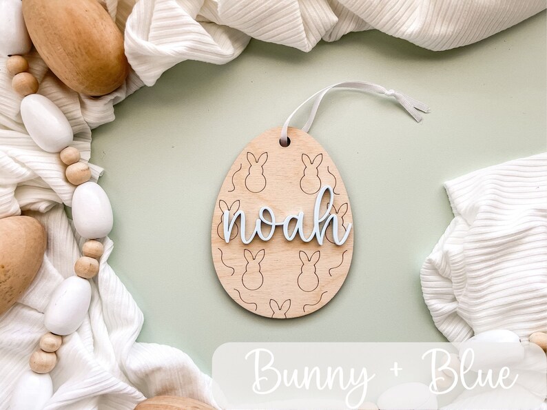 May include: A wooden Easter egg ornament with a bunny pattern and the name "noah" written in white. The ornament is hanging from a white ribbon and is surrounded by white fabric and wooden beads.