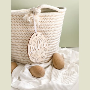 May include: A wooden Easter egg ornament with the name "Julie" and floral designs. The ornament is hanging from a white rope basket.