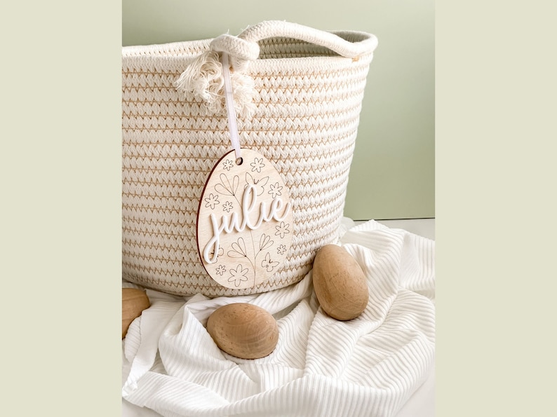 May include: A wooden Easter egg ornament with the name "Julie" and floral designs. The ornament is hanging from a white rope basket.