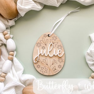 May include: Wooden Easter egg ornament with a butterfly and floral design. The egg is painted white and has the name "Julie" written on it.