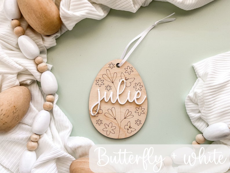 May include: Wooden Easter egg ornament with a butterfly and floral design. The egg is painted white and has the name "Julie" written on it.