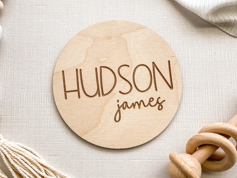 K&ouml;nnte beinhalten: Eine runde Holzplakette mit dem Namen "HUDSON james" in kursiv geschriebener Schrift.