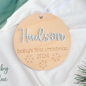 May include: A round wooden ornament with the name "Hudson" and the text "baby's first christmas 2024" in blue lettering. The ornament is decorated with snowflake designs.