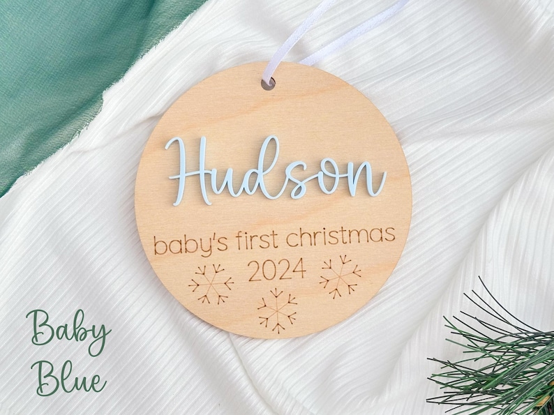 May include: A round wooden ornament with the name "Hudson" and the text "baby's first christmas 2024" in blue lettering. The ornament is decorated with snowflake designs.