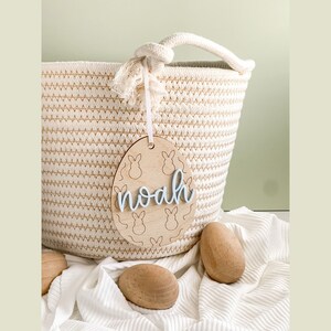 May include: A wooden Easter egg-shaped tag with bunny silhouettes and the name "noah" written in blue script. The tag is hanging from a woven basket.