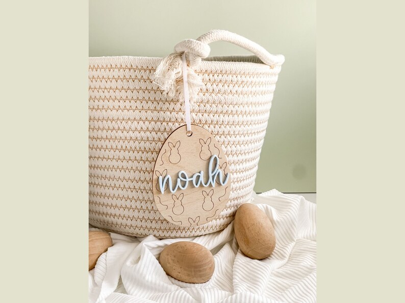 May include: A wooden Easter egg-shaped tag with bunny silhouettes and the name "noah" written in blue script. The tag is hanging from a woven basket.