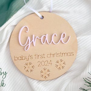 May include: A round wooden ornament with the name "Grace" in pink lettering, the text "baby's first christmas 2024" and three snowflake designs.