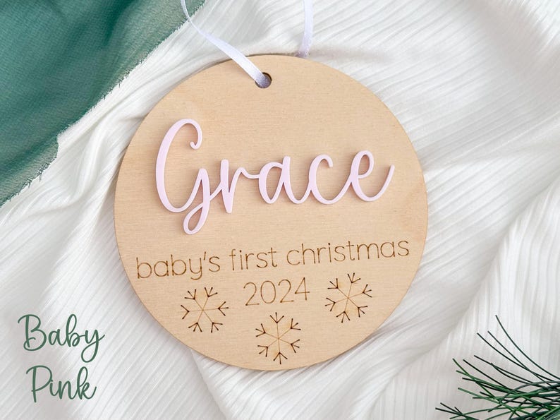 May include: A round wooden ornament with the name "Grace" in pink lettering, the text "baby's first christmas 2024" and three snowflake designs.