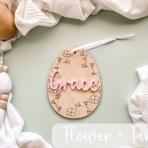 May include: A wooden egg-shaped ornament with a floral design and the name "Grace" in pink lettering. The ornament is hanging from a white ribbon.