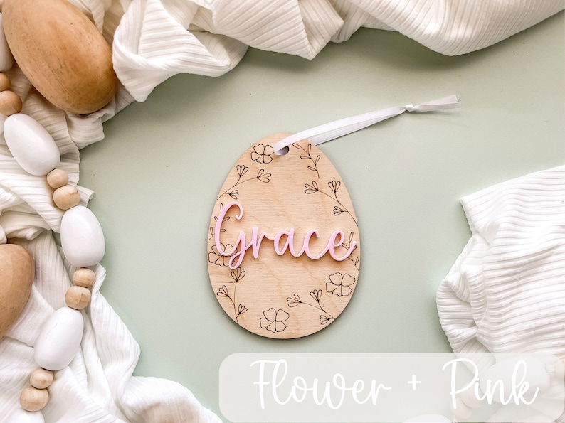 May include: A wooden egg-shaped ornament with a floral design and the name "Grace" in pink lettering. The ornament is hanging from a white ribbon.