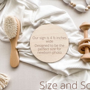 K&ouml;nnte beinhalten: Ein rundes Schild aus Holz mit dem Text "Our sign is 4 1/2 inches wide Designed to be the perfect size for newborn photo" auf einem wei&szlig;en Stoffhintergrund. Das Schild ist umgeben von einer Holzrassel, einer Holzhaarb&uuml;rste und einer wei&szlig;en Perlenkette.