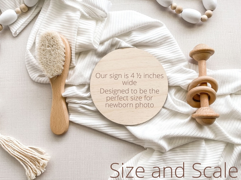 K&ouml;nnte beinhalten: Ein rundes Schild aus Holz mit dem Text "Our sign is 4 1/2 inches wide Designed to be the perfect size for newborn photo" auf einem wei&szlig;en Stoffhintergrund. Das Schild ist umgeben von einer Holzrassel, einer Holzhaarb&uuml;rste und einer wei&szlig;en Perlenkette.