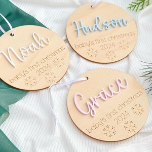 May include: Three wooden ornaments with white string hangers. Each ornament has a different name, "Noah", "Hudson", and "Grace", written in white raised letters. Each ornament also has the text "baby's first christmas" and the year "2024" engraved on the wood.  The ornaments are decorated with snowflake designs.