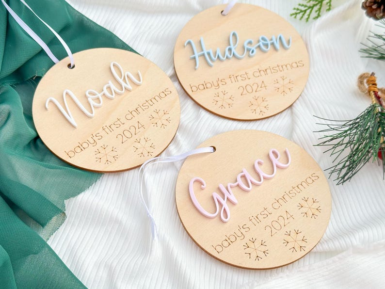 May include: Three wooden ornaments with white string hangers. Each ornament has a different name, "Noah", "Hudson", and "Grace", written in white raised letters. Each ornament also has the text "baby's first christmas" and the year "2024" engraved on the wood.  The ornaments are decorated with snowflake designs.