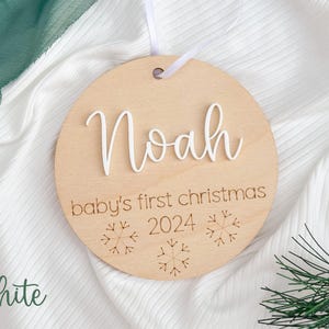 May include: White wooden ornament with the name "Noah" and the text "baby's first christmas 2024" with snowflake designs.