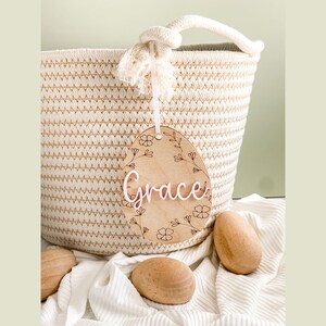 May include: A wooden egg-shaped ornament with a floral design and the name "Grace" written in white. The ornament is hanging from a white rope and is in front of a woven basket.