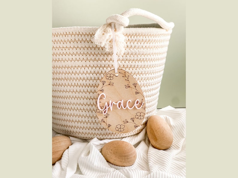 May include: A wooden egg-shaped ornament with a floral design and the name "Grace" written in white. The ornament is hanging from a white rope and is in front of a woven basket.