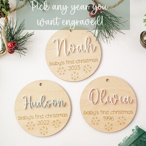 May include: Three round wooden ornaments with white lettering. The ornaments are decorated with snowflakes and the text "baby's first christmas". The ornaments are personalized with the names "Noah", "Hudson", and "Olivia" and the years "2023", "2022", and "1996".