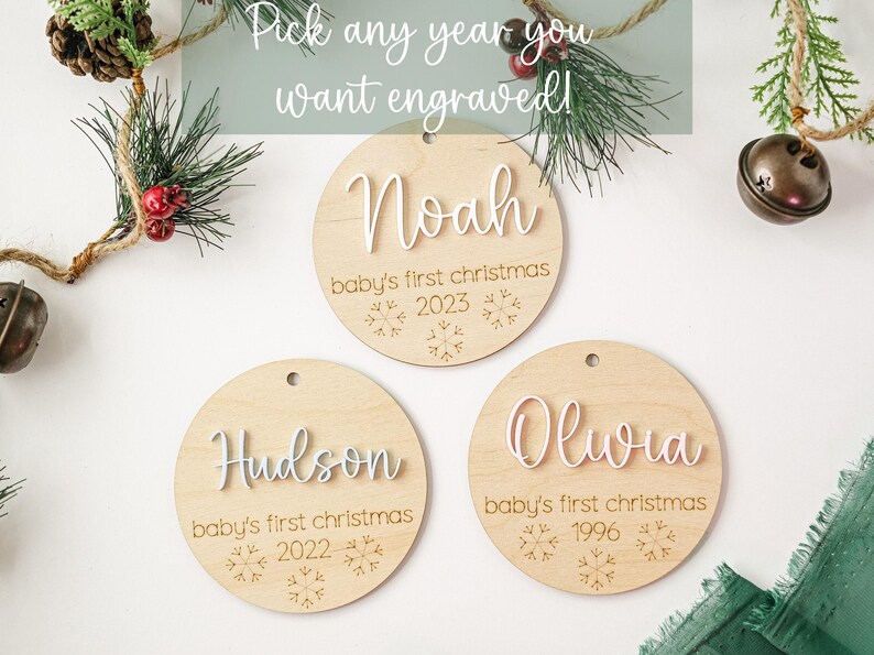 May include: Three round wooden ornaments with white lettering. The ornaments are decorated with snowflakes and the text "baby's first christmas". The ornaments are personalized with the names "Noah", "Hudson", and "Olivia" and the years "2023", "2022", and "1996".