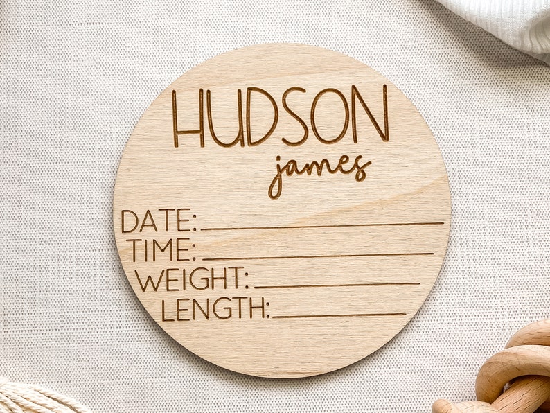 K&ouml;nnte beinhalten: Eine runde Holzplakette mit dem Namen "HUDSON james" eingraviert. Die Plakette hat auch die W&ouml;rter "DATE:", "TIME:", "WEIGHT:" und "LENGTH:" eingraviert, mit Linien zum Eintragen der Geburtsdaten.