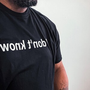 May include: A black short-sleeved t-shirt featuring the text "I don't know" printed in white capital letters across the chest. The text is oriented upside down relative to the neckline, creating a distinct visual contrast against the fabric.