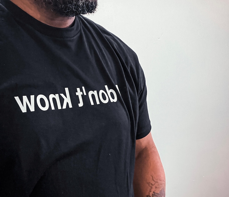 May include: A black short-sleeved t-shirt featuring the text "I don't know" printed in white capital letters across the chest. The text is oriented upside down relative to the neckline, creating a distinct visual contrast against the fabric.