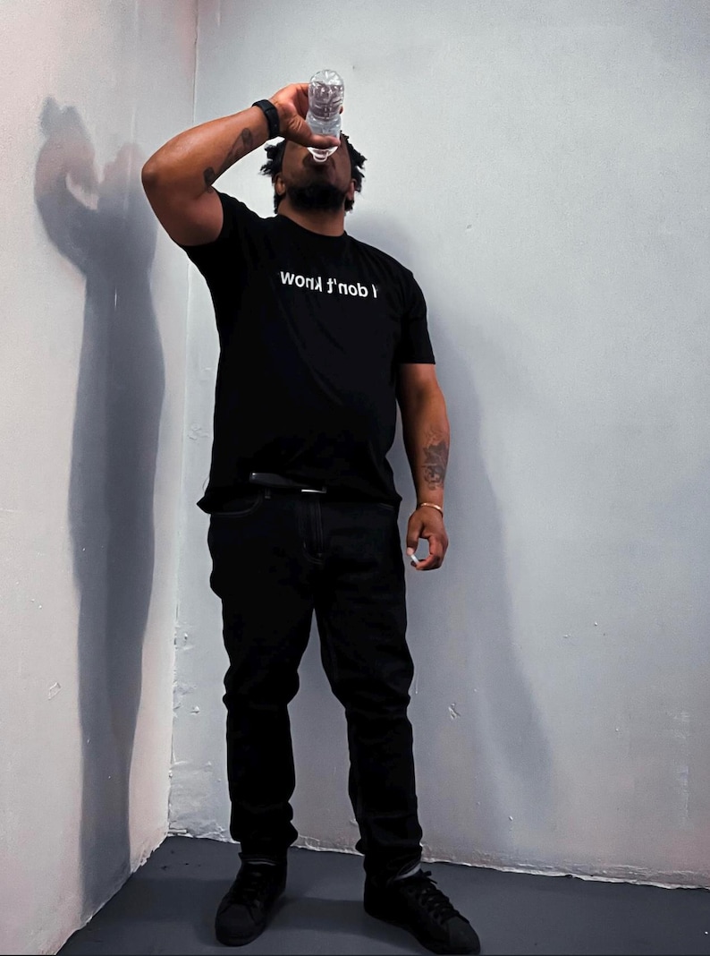 May include: A black short-sleeved t-shirt featuring the white text "I don't know" printed across the chest. The shirt is paired with black pants and black sneakers, creating a monochromatic look. The garment is worn by an individual drinking water.