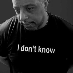 May include: A black short-sleeved t-shirt featuring the white text "I don't know" printed across the chest. The garment is worn by an individual with a short haircut and a thin chain necklace, captured in a black and white photographic style.