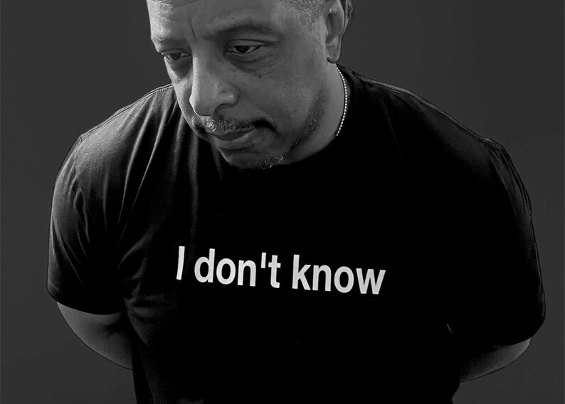 May include: A black short-sleeved t-shirt featuring the white text "I don't know" printed across the chest. The garment is worn by an individual with a short haircut and a thin chain necklace, captured in a black and white photographic style.
