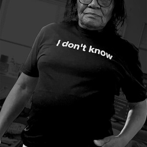 May include: A black short-sleeved t-shirt featuring the white text "I don't know" printed across the chest. The shirt is worn with white pants. The overall image is presented in a high-contrast black and white monochromatic color scheme.