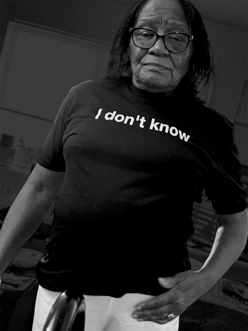May include: A black short-sleeved t-shirt featuring the white text "I don't know" printed across the chest. The shirt is worn with white pants. The overall image is presented in a high-contrast black and white monochromatic color scheme.
