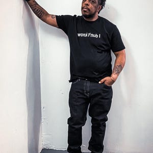 May include: A black short-sleeved t-shirt featuring the white text "I don't know" printed in a mirrored, reversed orientation across the chest. The shirt is paired with dark-colored denim jeans and black sneakers, creating a monochromatic look.