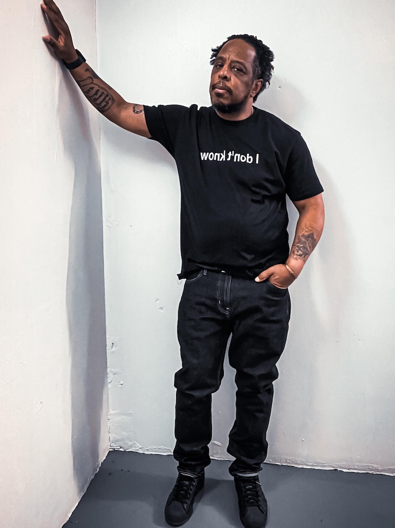 May include: A black short-sleeved t-shirt featuring the white text "I don't know" printed in a mirrored, reversed orientation across the chest. The shirt is paired with dark-colored denim jeans and black sneakers, creating a monochromatic look.