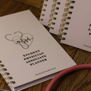 Könnte beinhalten: Ein rosa Spiralblock mit dem Text "Student Physician Associate Planner" und dem Hashtag "#ladiesinhealthcare". Ein Stethoskop liegt auf einer Holzoberfläche neben dem Block.
