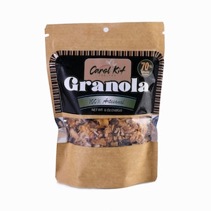 Puede incluir: Una bolsa de papel marr&oacute;n con una ventana de pl&aacute;stico transparente que muestra granola en su interior. La bolsa tiene una etiqueta negra con el texto "Carol Kit HEALTHY Granola 100% Artesanal NET WT 6 Oz (168Gr)" y un c&iacute;rculo rojo con el texto "70% Whole Grains".