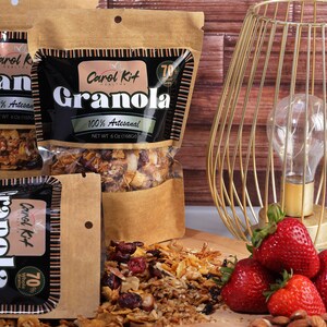Puede incluir: Tres bolsas de granola con el texto "Carol Kit Granola 100% Artesanal 70% Whole Grains" en la parte delantera. Las bolsas son marrones con texto negro y est&aacute;n sobre una superficie de madera con fresas y almendras.