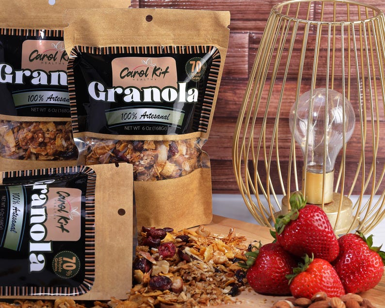 Puede incluir: Tres bolsas de granola con el texto "Carol Kit Granola 100% Artesanal 70% Whole Grains" en la parte delantera. Las bolsas son marrones con texto negro y est&aacute;n sobre una superficie de madera con fresas y almendras.