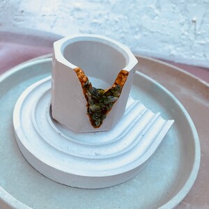 May include: A white, geometric concrete planter with a gold-filled crack, holding green stones. The planter sits on a matching white, tiered saucer. The design is minimalist and modern, suitable for small plants or succulents.