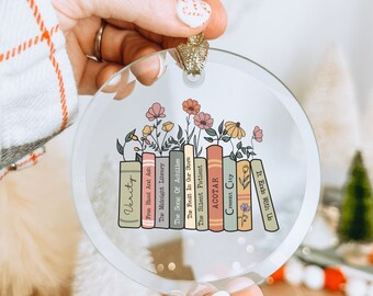 May include: A glass ornament featuring a stack of books with floral accents. The book titles include "Verity", "From Blood And Ash", "The Midnight Library", "The Song Of Achilles", "The Fault In Our Stars", "The Silent Patient", "ACOTAR", "Crescent City", and "It Ends With Us".